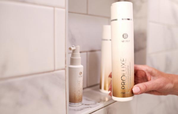 Image of ProLuxe Shampoo bottle held by hand moving it onto a shower shelf next to the rest of the ProLuxe Hair Care System.
